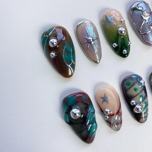 K&ouml;nnte beinhalten: Nahaufnahme von zehn verschiedenen Nagelkunstdesigns. Die Designs zeigen verschiedene Farben, Muster und Verzierungen, darunter silberne Sterne, gr&uuml;ne und braune Wirbel und silberne Perlen.