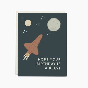 May include: A dark blue card with a brown space shuttle flying through space with two moons and stars. The text on the card reads "Hope your birthday is a blast."