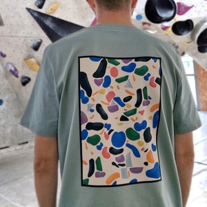 Op de afbeelding: Een lichtblauw T-shirt met een kleurrijk abstract ontwerp op de rug. Het ontwerp bevat een verscheidenheid aan vormen en kleuren, waaronder blauw, groen, oranje en roze.