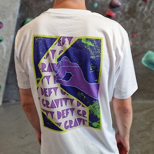 May include: White short-sleeved t-shirt with a graphic on the back. The graphic features a purple hand and the words "Defy Gravity" in a repeating pattern, set against a purple and blue background with a green border.