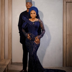 May include: A couple in traditional Nigerian attire. The woman is wearing a blue, beaded, mermaid-style gown with a matching head wrap. The man is wearing a black suit with a blue cap.