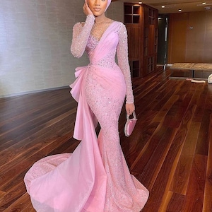 May include: A pink sequined gown with a long train and a sheer overlay. The gown has a fitted bodice and a mermaid silhouette. The model is wearing a pink headscarf.