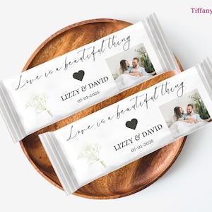 Könnte beinhalten: Zwei weiße Schokoriegelverpackungen mit dem Text "Love is a beautiful thing", einem schwarzen Herz, Namen und einem Datum. Jede Verpackung hat ein Foto eines Paares. Die Verpackungen liegen auf einem Holztablett.