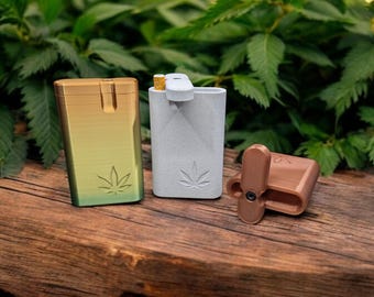 May include: Three small, rectangular cases in gold, gray, and brown, each with a cannabis leaf design. The gold case has a gradient finish. The gray case has a white mouthpiece. The brown case is open, revealing a small compartment. All three are on a wooden surface.