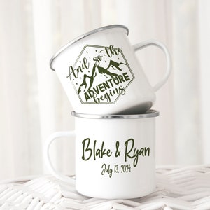 May include: Two white enamel mugs with black rims. The top mug features a green mountain design with the text "And so the adventure begins". The bottom mug has the text "Blake & Ryan July 13, 2024".