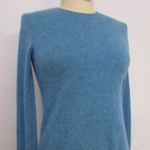May include: A light blue crew neck sweater with long sleeves. The sweater is displayed on a mannequin. The fabric has a soft, textured appearance. The mannequin has text that reads "COLLAPSIBLE FABULOUS FIT 2005 PRO SERIES".