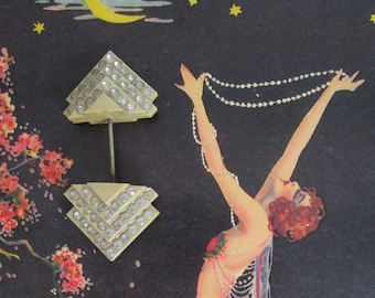 May include: A pair of vintage, white, geometric, rhinestone brooches. Each brooch is shaped like a triangle with a pointed top and a flat base. The brooches are adorned with clear rhinestones that are set in a geometric pattern. The brooches are attached to a metal pin.