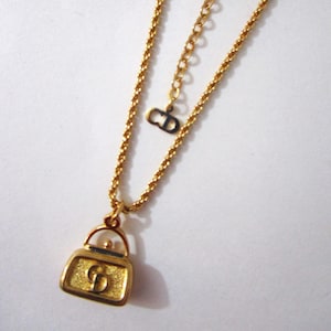May include: A gold-tone necklace with a small handbag charm. The charm has a textured surface and the letters "CD" are visible on the front. The necklace has a lobster clasp closure.