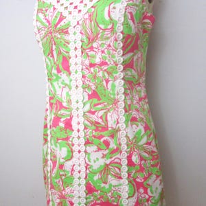 May include: A sleeveless dress with a vibrant floral pattern in pink, green, and white. The dress features a white crochet neckline and trim, creating a contrast against the colorful print. The dress is displayed on a mannequin.
