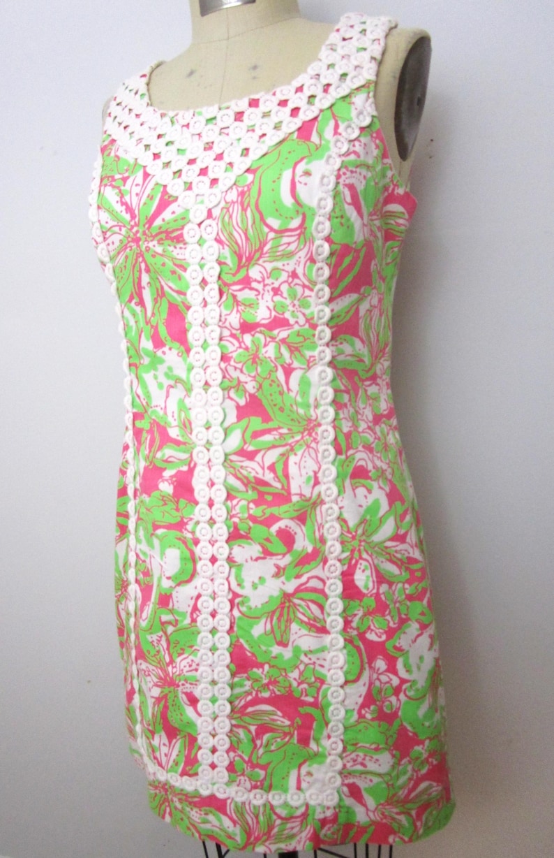 May include: A sleeveless dress with a vibrant floral pattern in pink, green, and white. The dress features a white crochet neckline and trim, creating a contrast against the colorful print. The dress is displayed on a mannequin.