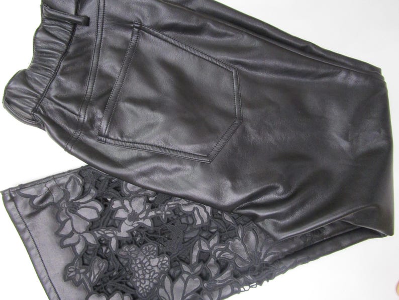 Pu&ograve; includere: Pantaloni neri con finitura liscia e una tasca posteriore. La parte inferiore della gamba presenta un intricato motivo floreale in pizzo nero intagliato che si estende sul tessuto, creando un contrasto dettagliato con il materiale nero solido.