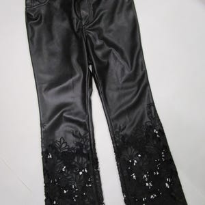 Pu&ograve; includere: Pantaloni neri con una finitura liscia simile alla pelle, dotati di chiusura con bottone e cerniera, tasche anteriori e intricati dettagli in pizzo floreale nero intagliato sul fondo di ogni gamba, che risalgono dall'orlo.