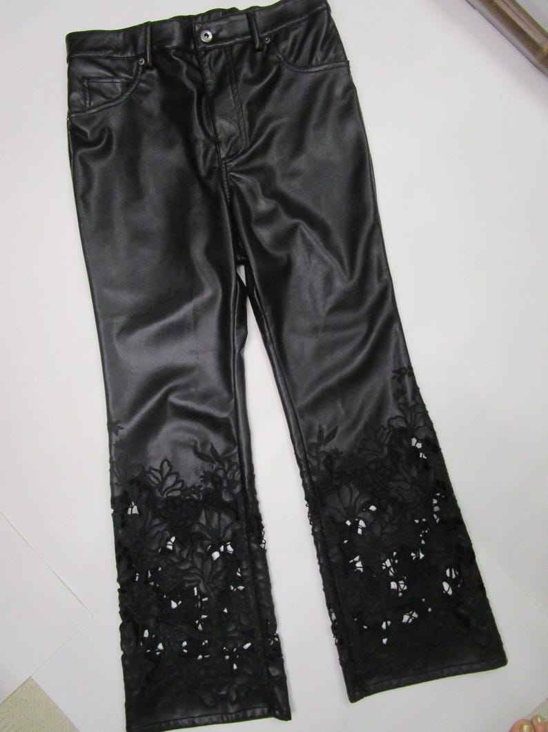 Pu&ograve; includere: Pantaloni neri con una finitura liscia simile alla pelle, dotati di chiusura con bottone e cerniera, tasche anteriori e intricati dettagli in pizzo floreale nero intagliato sul fondo di ogni gamba, che risalgono dall'orlo.