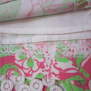May include: Close-up of a folded fabric with a floral pattern in pink, green, and white. The fabric has a white lining and a white lace trim. The bottom edge features a white, circular, crocheted trim.