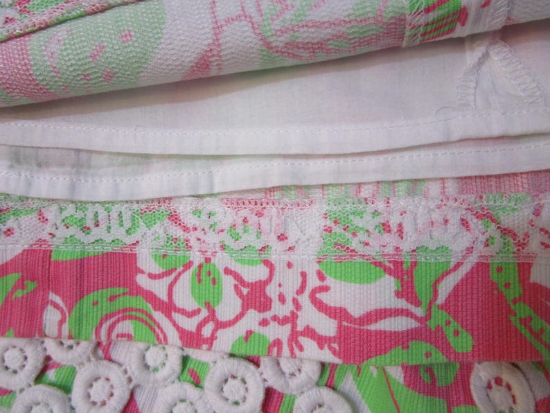 May include: Close-up of a folded fabric with a floral pattern in pink, green, and white. The fabric has a white lining and a white lace trim. The bottom edge features a white, circular, crocheted trim.