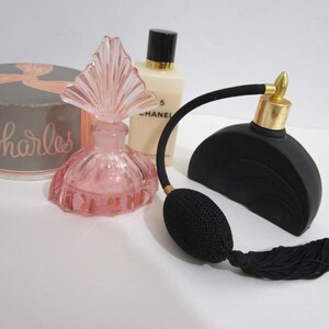 Pode incluir: Três frascos de perfume vintage. Um frasco de vidro rosa com uma tampa decorativa, um frasco de vidro preto com uma tampa dourada e uma borla preta, e um frasco branco com uma tampa preta e o texto "Chanel No. 5" no rótulo.