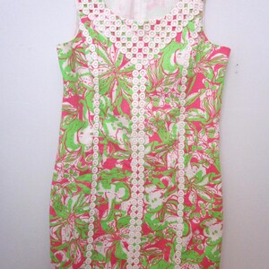 May include: Sleeveless dress with a floral pattern in pink, green, and white. The dress features a white lace trim around the neckline and down the front. The dress is hanging on a pink hanger.