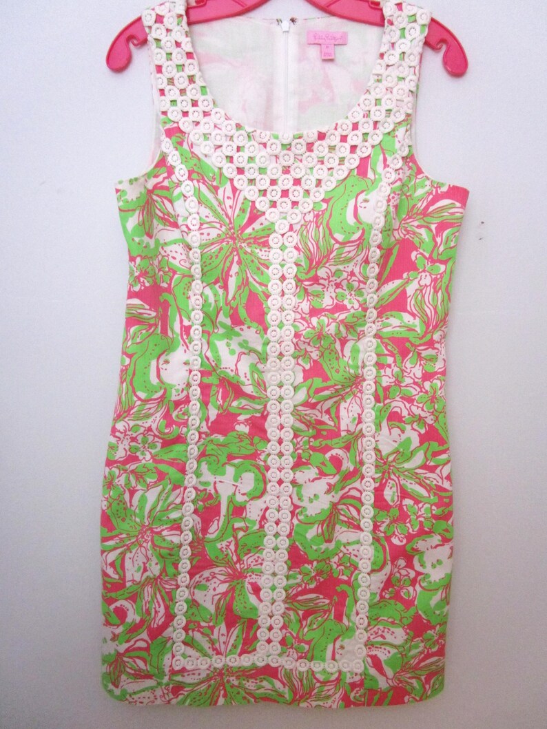 May include: Sleeveless dress with a floral pattern in pink, green, and white. The dress features a white lace trim around the neckline and down the front. The dress is hanging on a pink hanger.
