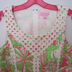 May include: A sleeveless dress with a floral pattern in pink and green, accented with white lace trim around the neckline. The dress is hanging on a pink hanger.