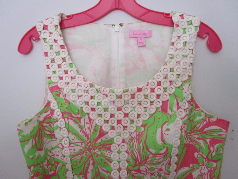 May include: A sleeveless dress with a floral pattern in pink and green, accented with white lace trim around the neckline. The dress is hanging on a pink hanger.