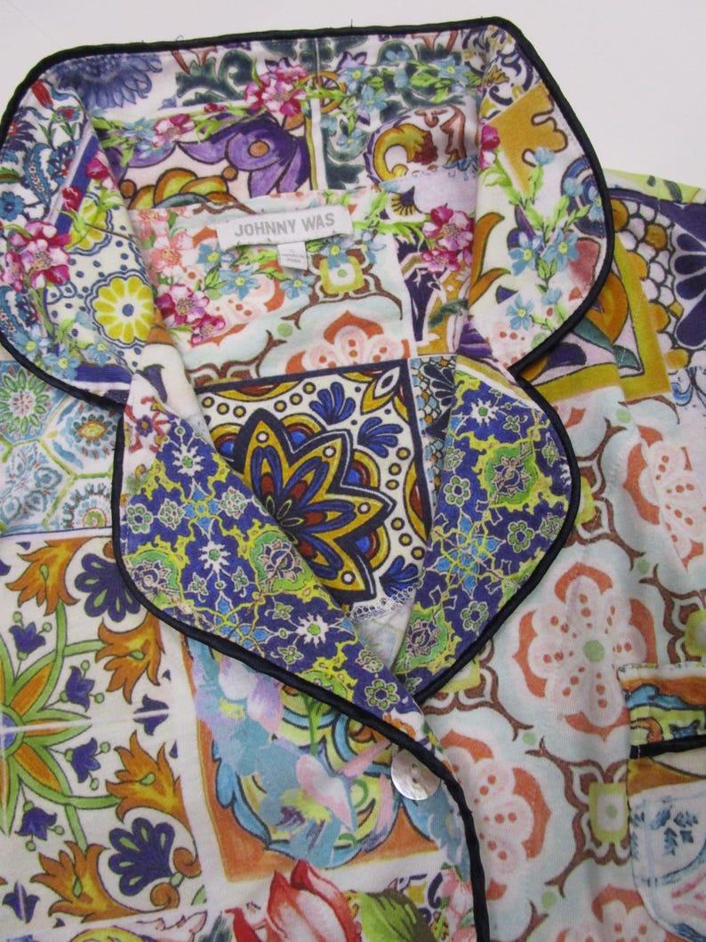 Puede incluir: Una camisa abotonada con cuello de solapa y ribete negro, con un estampado multicolor inspirado en azulejos con motivos florales y geom&eacute;tricos en tonos azul, amarillo, naranja y verde. Una etiqueta blanca muestra el texto JOHNNY WAS.