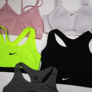 May include: Assortment of Nike sports bras in various colors, including pink, white, neon green, black, and gray. Each bra features the Nike swoosh logo. Styles include racerback and adjustable strap designs.