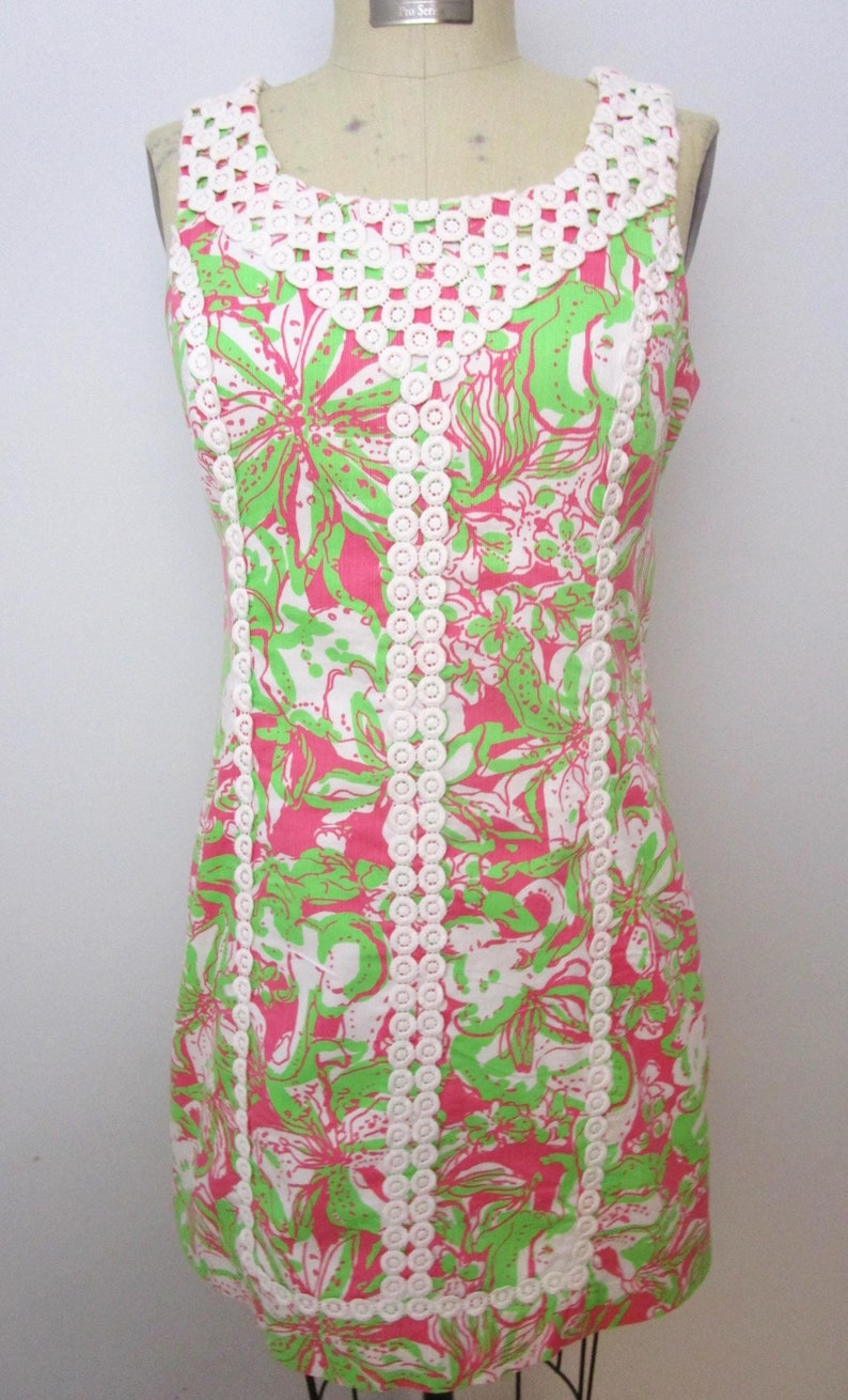 May include: A sleeveless dress with a floral pattern in pink, green, and white. The dress features a white crocheted neckline and trim. The dress is displayed on a mannequin.