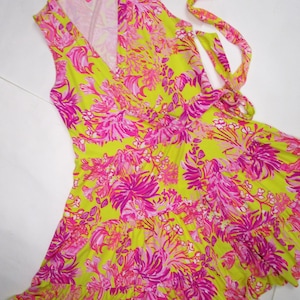 May include: A sleeveless wrap dress featuring a vibrant floral pattern in shades of pink and fuchsia on a bright yellow-green background. The dress has a V-neckline and a flowing, ruffled hemline, with tie closures at the waist.