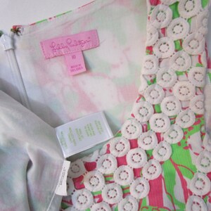 May include: A sleeveless dress with a vibrant floral pattern in pink and green. The dress features a white lace trim around the neckline and a zipper closure. The tag reads "Lilly Pulitzer" and "10".