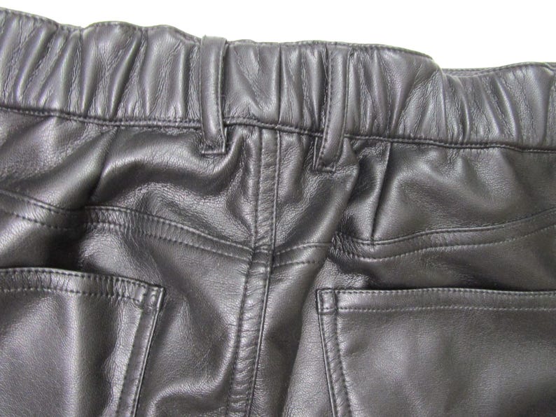 Pu&ograve; includere: Pantaloni in pelle nera con vita elasticizzata e passanti integrati, dettagli con impunture a vista e due tasche posteriori. Il materiale presenta una finitura liscia e leggermente testurizzata con motivi a grana naturale sulla superficie.