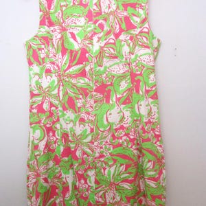 May include: Sleeveless dress with a vibrant floral pattern in pink, white, and green. The dress is hanging on a pink hanger, showcasing its colorful design. The floral print is a repeating pattern.