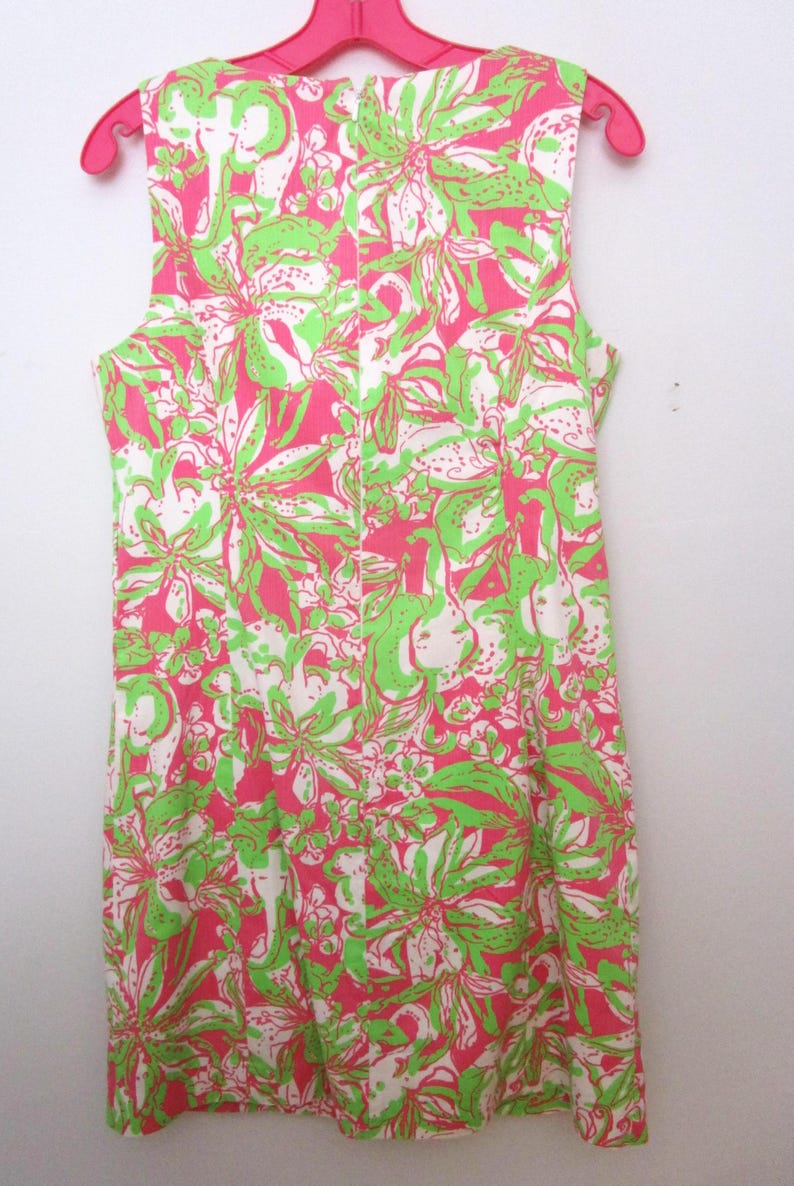 May include: Sleeveless dress with a vibrant floral pattern in pink, white, and green. The dress is hanging on a pink hanger, showcasing its colorful design. The floral print is a repeating pattern.