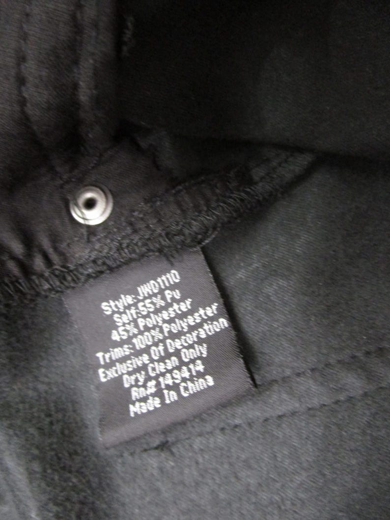 Pu&ograve; includere: L'interno di un capo nero con un'etichetta di manutenzione cucita che riporta il testo bianco: Style: JWD1110, Self: 55% Pu, 45% Polyester, Trims: 100% Polyester, Exclusive Of Decoration, Dry Clean Only, Rn# 149414, Made In China.