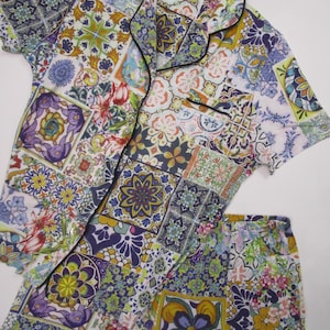 May include: A short-sleeve button-down pajama shirt and matching shorts set featuring a vibrant, multi-colored tile pattern. The fabric displays a patchwork of floral and geometric motifs in shades of purple, yellow, green, and blue, finished with a black piped trim along the collar and front pocket.