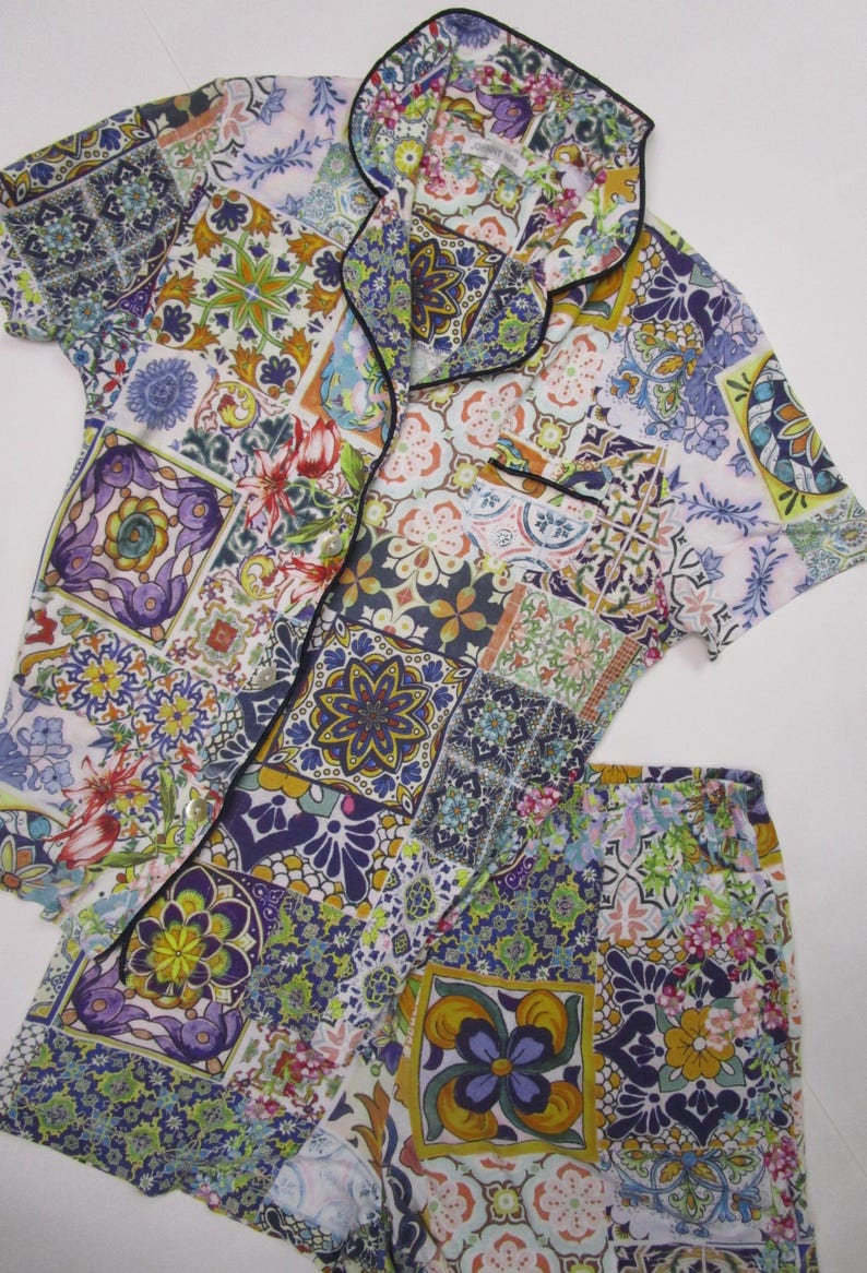Puede incluir: Un conjunto de pijama de camisa de manga corta con botones y pantal&oacute;n corto a juego, con un vibrante patr&oacute;n de azulejos multicolor. La tela muestra un mosaico de motivos florales y geom&eacute;tricos en tonos morado, amarillo, verde y azul, con un ribete negro en el cuello y el bolsillo delantero.