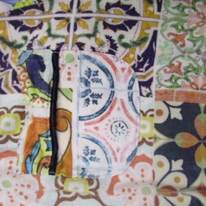 Puede incluir: Una prenda de tela con un dise&ntilde;o de patchwork de varios patrones de azulejos de estilo mediterr&aacute;neo en tonos azul, verde, naranja y amarillo. La pieza incluye un bolsillo redondeado con un borde vertical oscuro y un peque&ntilde;o bot&oacute;n verde.