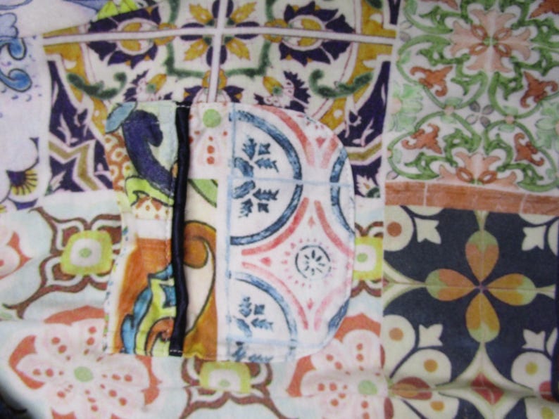 Puede incluir: Una prenda de tela con un dise&ntilde;o de patchwork de varios patrones de azulejos de estilo mediterr&aacute;neo en tonos azul, verde, naranja y amarillo. La pieza incluye un bolsillo redondeado con un borde vertical oscuro y un peque&ntilde;o bot&oacute;n verde.