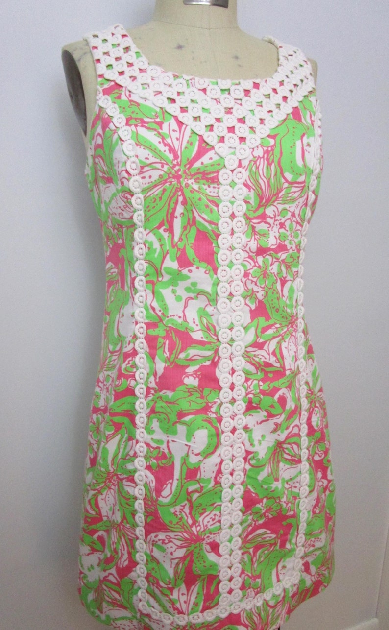 May include: Sleeveless dress with a floral pattern in pink, green, and white. The dress features a white crochet neckline and trim. The dress is displayed on a mannequin.