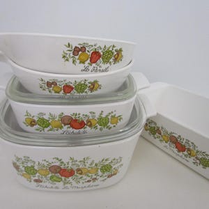 May include: A collection of white ceramic cookware with a colorful vegetable and herb design. The set includes a rectangular baking dish and several lidded and handled dishes. The text "Le Persil", "L'Echalote", and "La Marjolaine" are printed on the dishes.