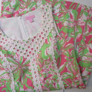 May include: A sleeveless dress with a floral pattern in pink, green, and white. The dress features a white, openwork trim around the neckline and armholes. A white zipper runs down the front. The dress is folded, showing the pattern on the fabric.
