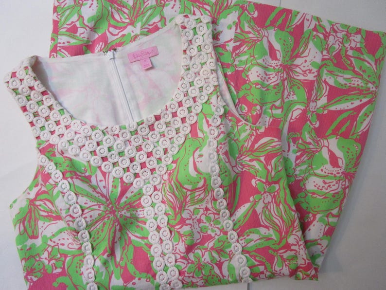 May include: A sleeveless dress with a floral pattern in pink, green, and white. The dress features a white, openwork trim around the neckline and armholes. A white zipper runs down the front. The dress is folded, showing the pattern on the fabric.