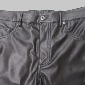 Pu&ograve; includere: Pantaloni in pelle nera con un classico design a cinque tasche, chiusura frontale con bottone, patta con cerniera e passanti per cintura. Il materiale liscio ha una leggera lucentezza, con cuciture visibili lungo le tasche e la vita.