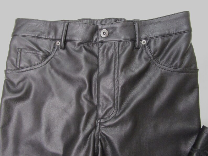 Pu&ograve; includere: Pantaloni in pelle nera con un classico design a cinque tasche, chiusura frontale con bottone, patta con cerniera e passanti per cintura. Il materiale liscio ha una leggera lucentezza, con cuciture visibili lungo le tasche e la vita.