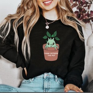 May include: Black sweatshirt featuring a cartoon plant creature in a terracotta pot with the text "THINK OUTSIDE THE POT". The creature is green with large eyes and is surrounded by green leaves. The sweatshirt is worn by a person.
