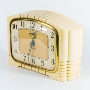 May include: Cream-colored vintage alarm clock with a gold-tone frame and a tan face. The clock features black numerals and hands, with a small seconds dial. The clock has a rectangular shape with rounded edges and a ribbed design on the side.