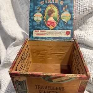 May include: An open wooden cigar box with a hinged lid featuring a portrait of a woman in a red dress. The lid text reads DIXIE'S and DIXIE CIGARS. The front of the box is labeled TRAVELLERS SELECTOS, with additional text printed on the interior sides.