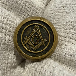 May include: A circular brass object featuring a raised Masonic square and compass emblem with a central letter G. The emblem is set against a dark, textured background within the brass frame, which has a polished, metallic finish and a rounded edge.