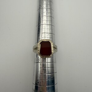 May include: A silver ring with a rectangular, reddish-brown stone. The ring is set on a silver ring sizer. The ring has a decorative design on the band. The stone is a deep, rich color, and the ring appears to be vintage.