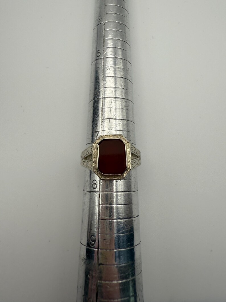 May include: A silver ring with a rectangular, reddish-brown stone. The ring is set on a silver ring sizer. The ring has a decorative design on the band. The stone is a deep, rich color, and the ring appears to be vintage.
