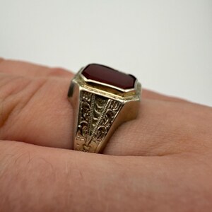 May include: A silver ring with a rectangular, deep red gemstone. The ring has an ornate, engraved design on the band. The gemstone is set in a gold-colored bezel. The ring is shown on a person's finger.